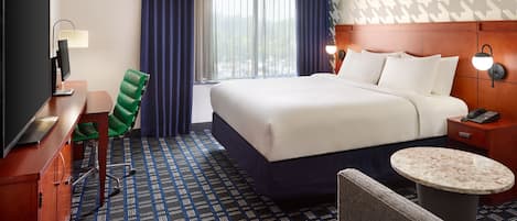 A hotel room with a bed, a desk with a computer, a chair, a small table, and a nightstand with a lamp.