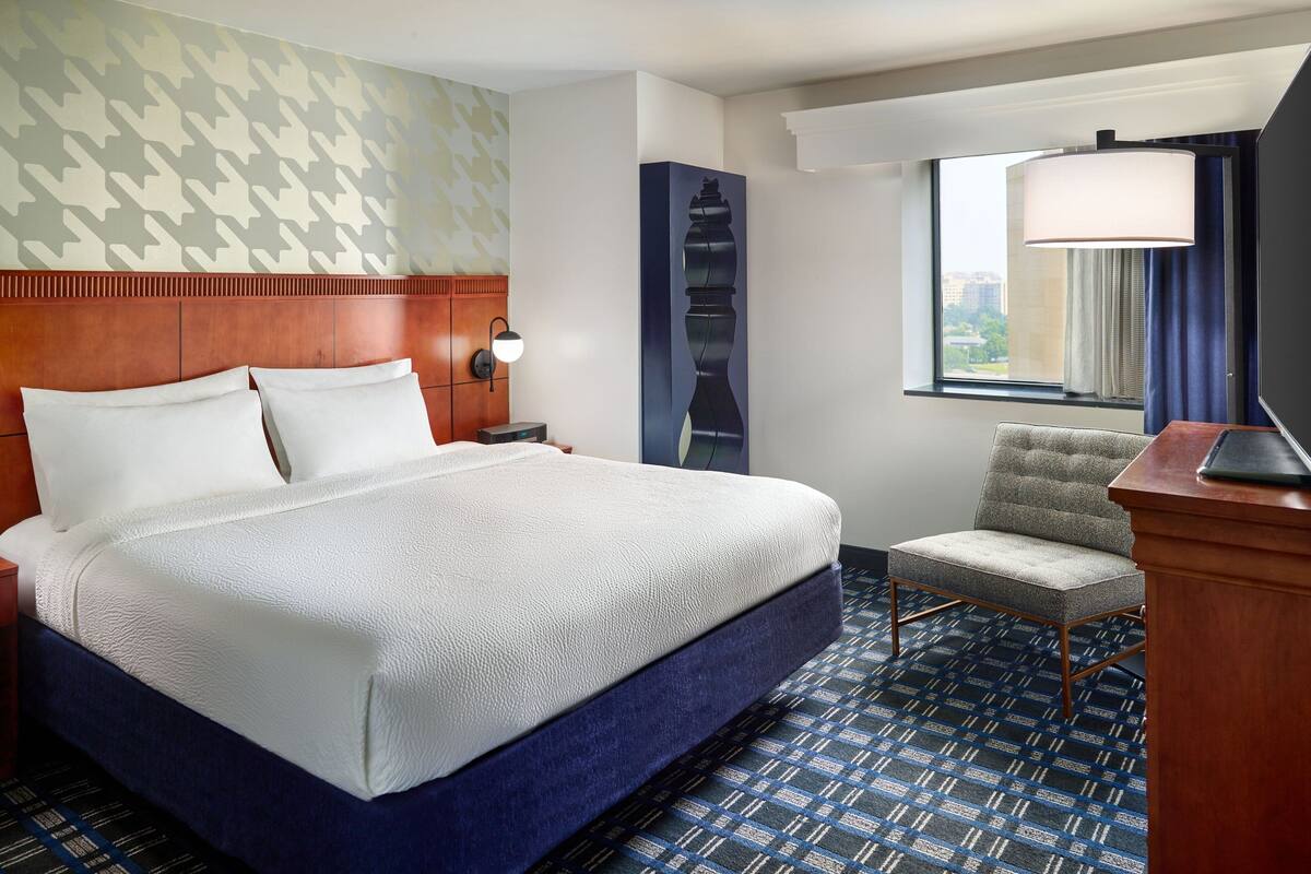 Une chambre d’hôtel avec un grand lit, une chaise, un bureau, une lampe et une fenêtre donnant sur la ville.