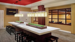 Un moderno hall de hotel con un mostrador de recepción alargado, varios taburetes de bar, un amplio ventanal con cortinas y un televisor montado en la pared.