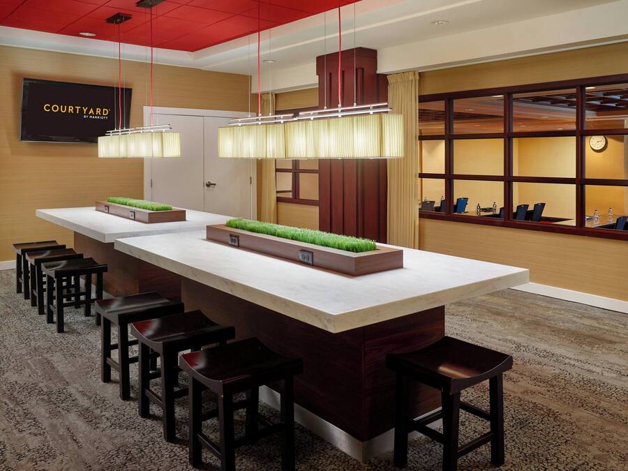 A modern hotel lobby with a long reception desk, multiple bar stools, a large window with curtains, and a television mounted on the wall.