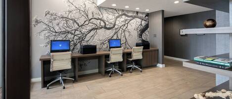 Um espaço de escritório moderno com três mesas, cada uma com monitor de computador, cadeiras de couro e um mural grande na parede com galhos de árvores.