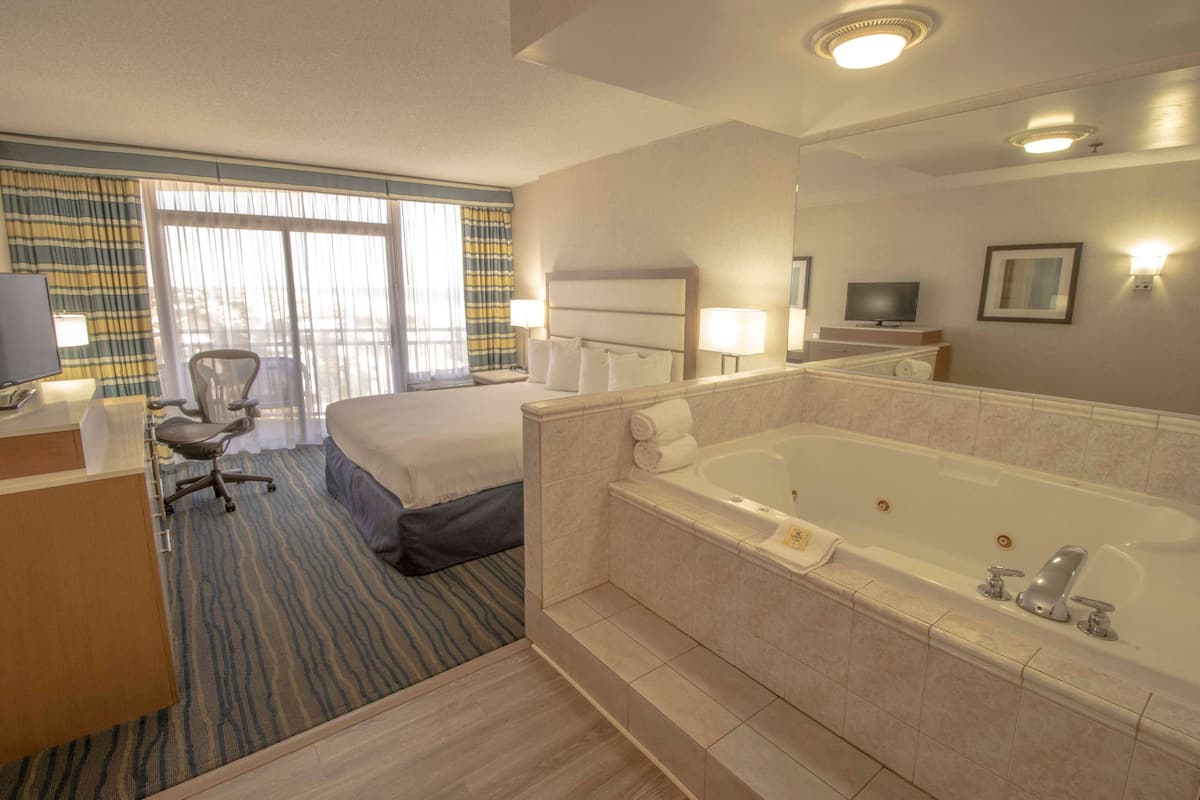 Habitación de hotel con una cama grande, una zona de estar separada con una silla, un escritorio, un televisor, una bañera amplia y una ventana con cortinas.