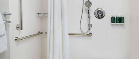 A shower area with a white curtain, grab bars, a handheld showerhead, and a small table with a towel.