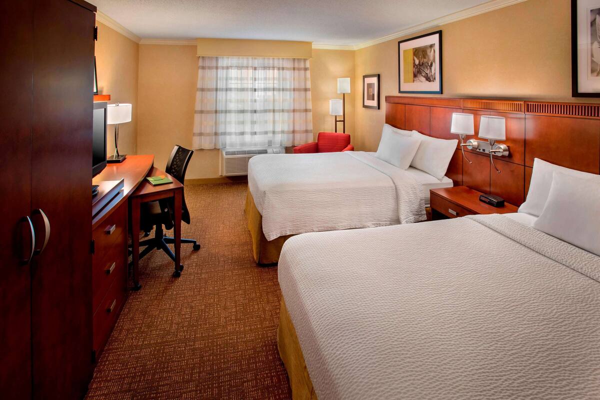 A hotel room with two beds, a desk, a chair, a lamp, and a window with curtains.