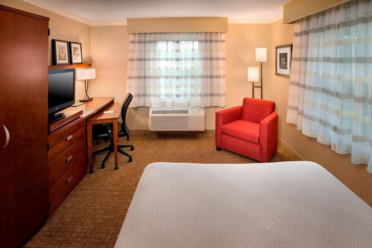 A hotel room with a bed, a desk with a computer, a red chair, a TV, and a window with curtains.