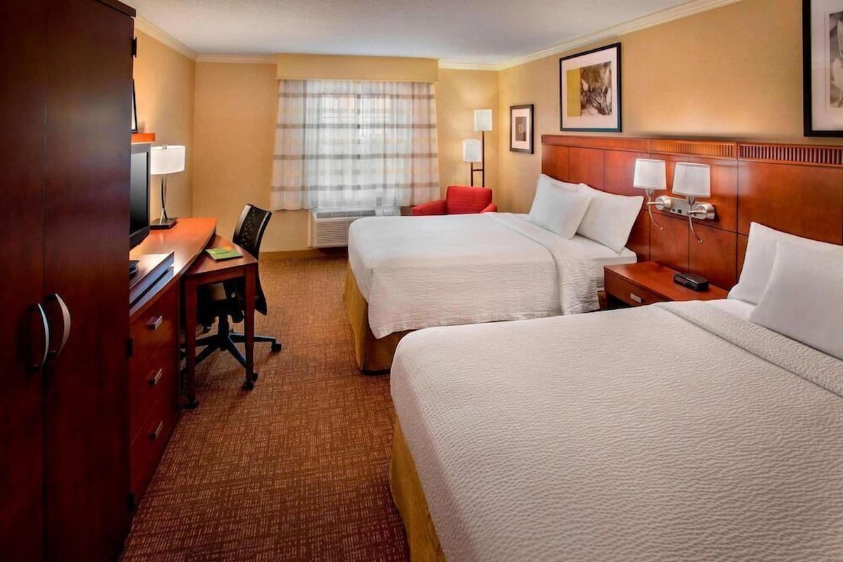 A hotel room with two beds, a desk with a chair, a lamp, and a window with curtains.
