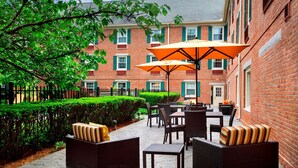 An outdoor patio with wicker furniture and orange umbrellas, surrounded by a brick building and greenery. - Sonesta Select Boston Danvers (Danvers)