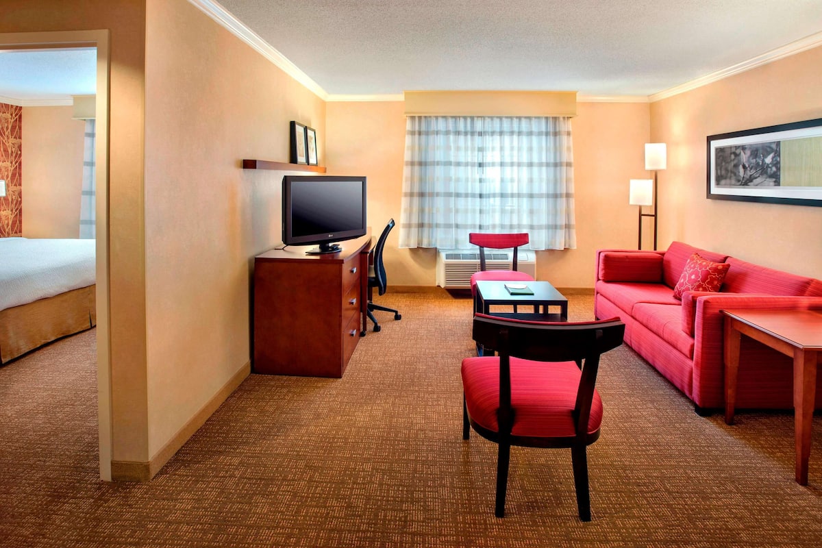 A hotel room with a bed, a desk with a computer, a red sofa, a red chair, and a window with curtains.
