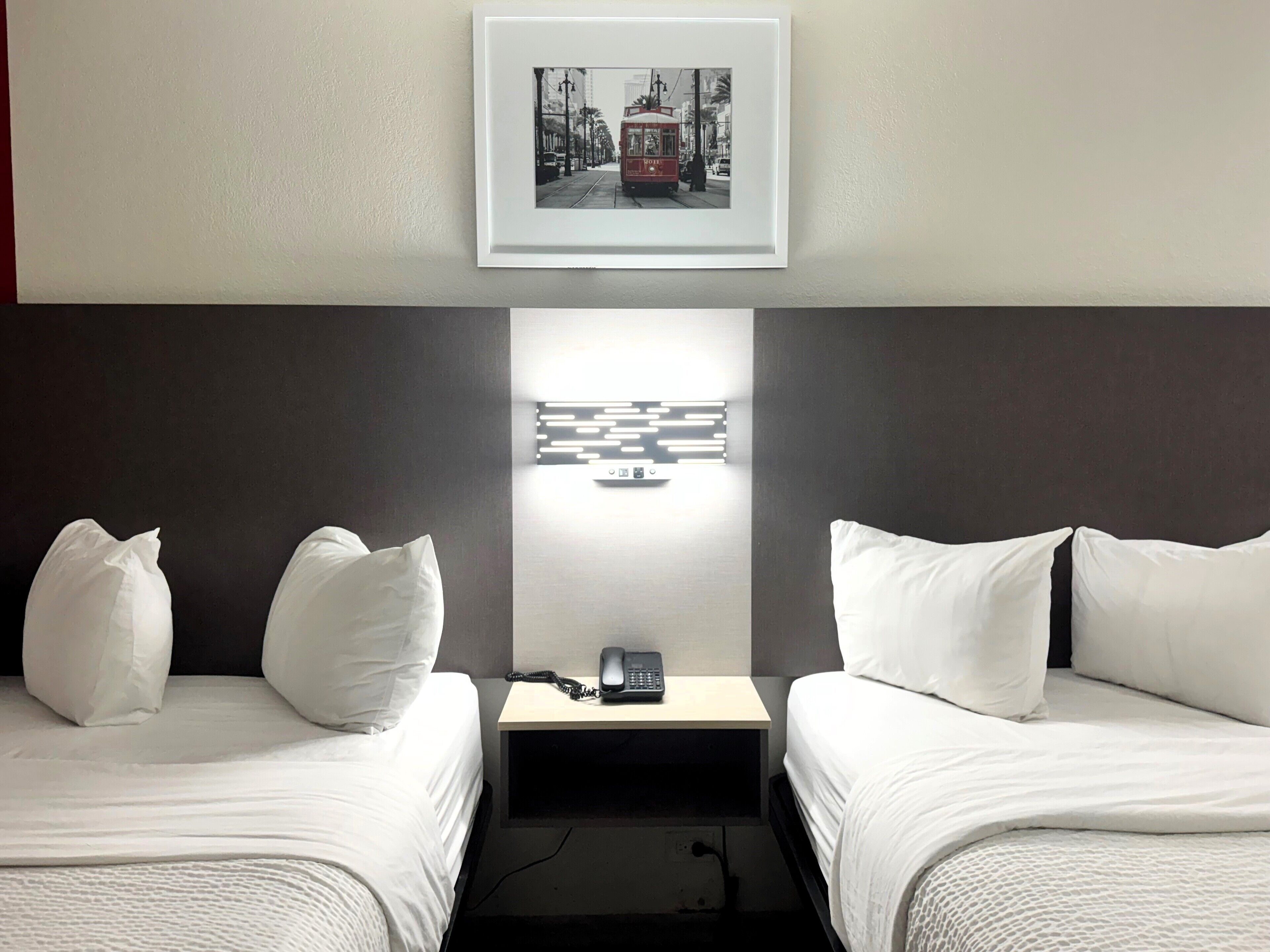Une chambre d’hôtel avec deux lits, une table de chevet avec un téléphone, une lampe fixée au mur et un tableau encadré au-dessus du lit.