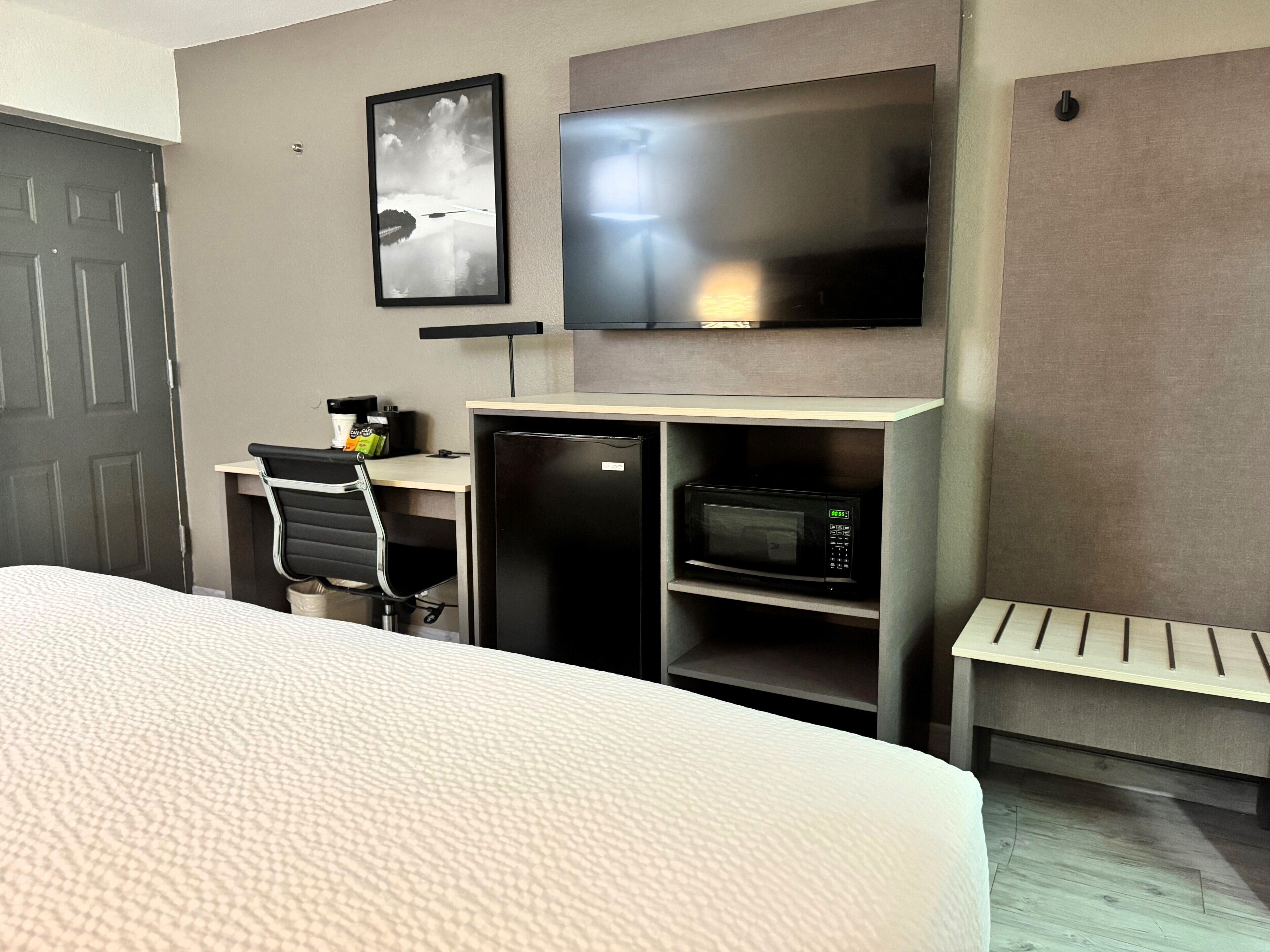 A hotel room with a bed, a desk with a chair, a television, a microwave, and a small shelf.