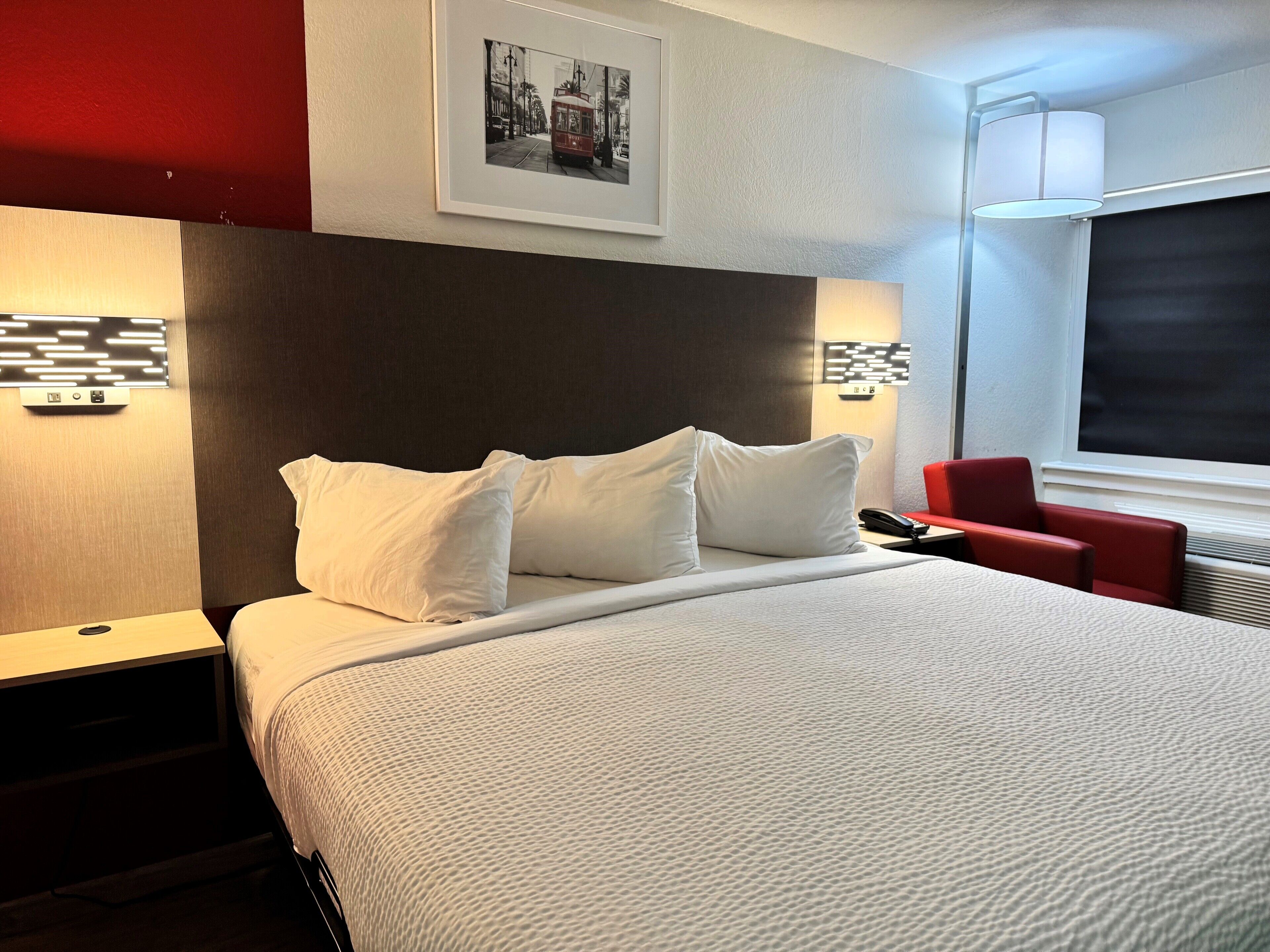 A hotel room with a bed, white pillows, a red chair, a framed photo on the wall, and a window with a view.