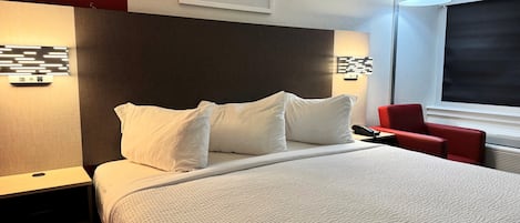 A hotel room with a bed, white pillows, a red chair, a framed photo on the wall, and a window with a view.