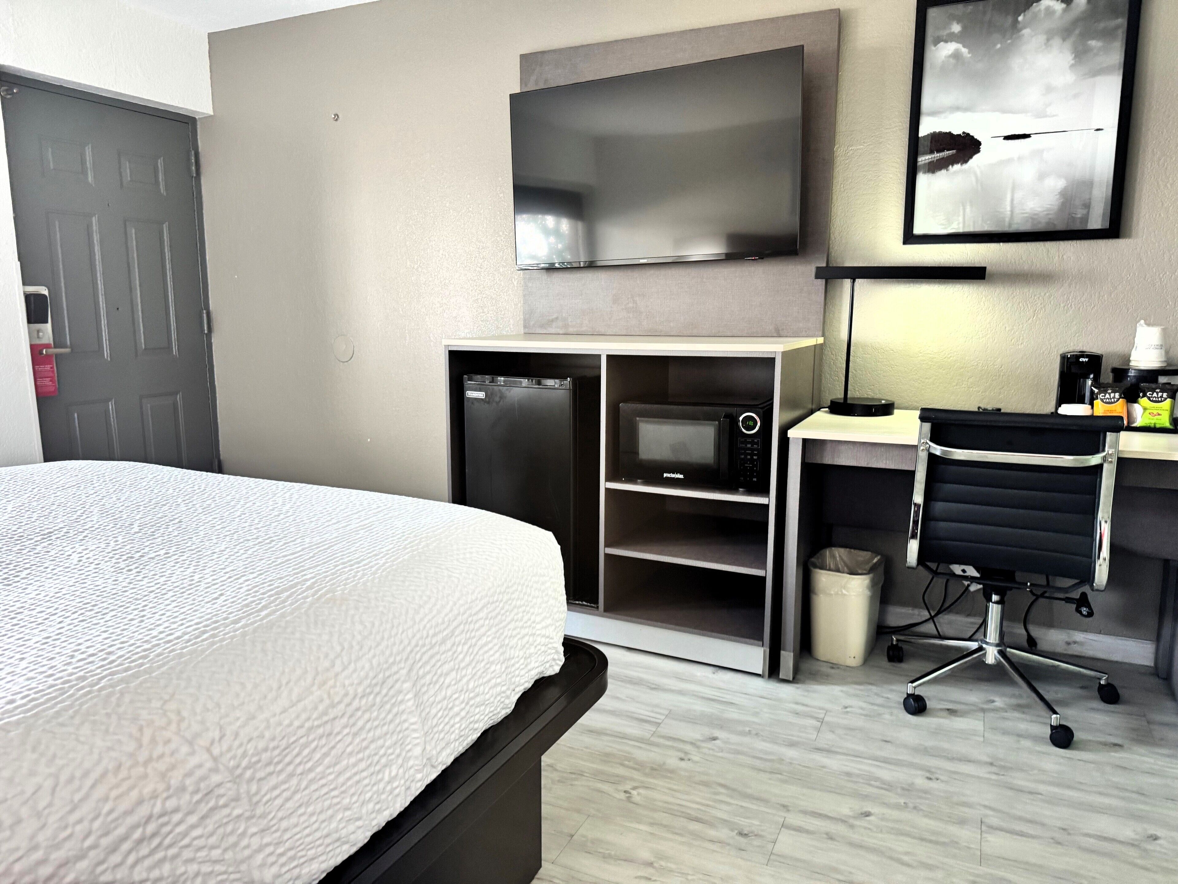 Una habitación de hotel con una cama, un escritorio con silla, un microondas, un cesto de basura y un televisor montado en la pared.