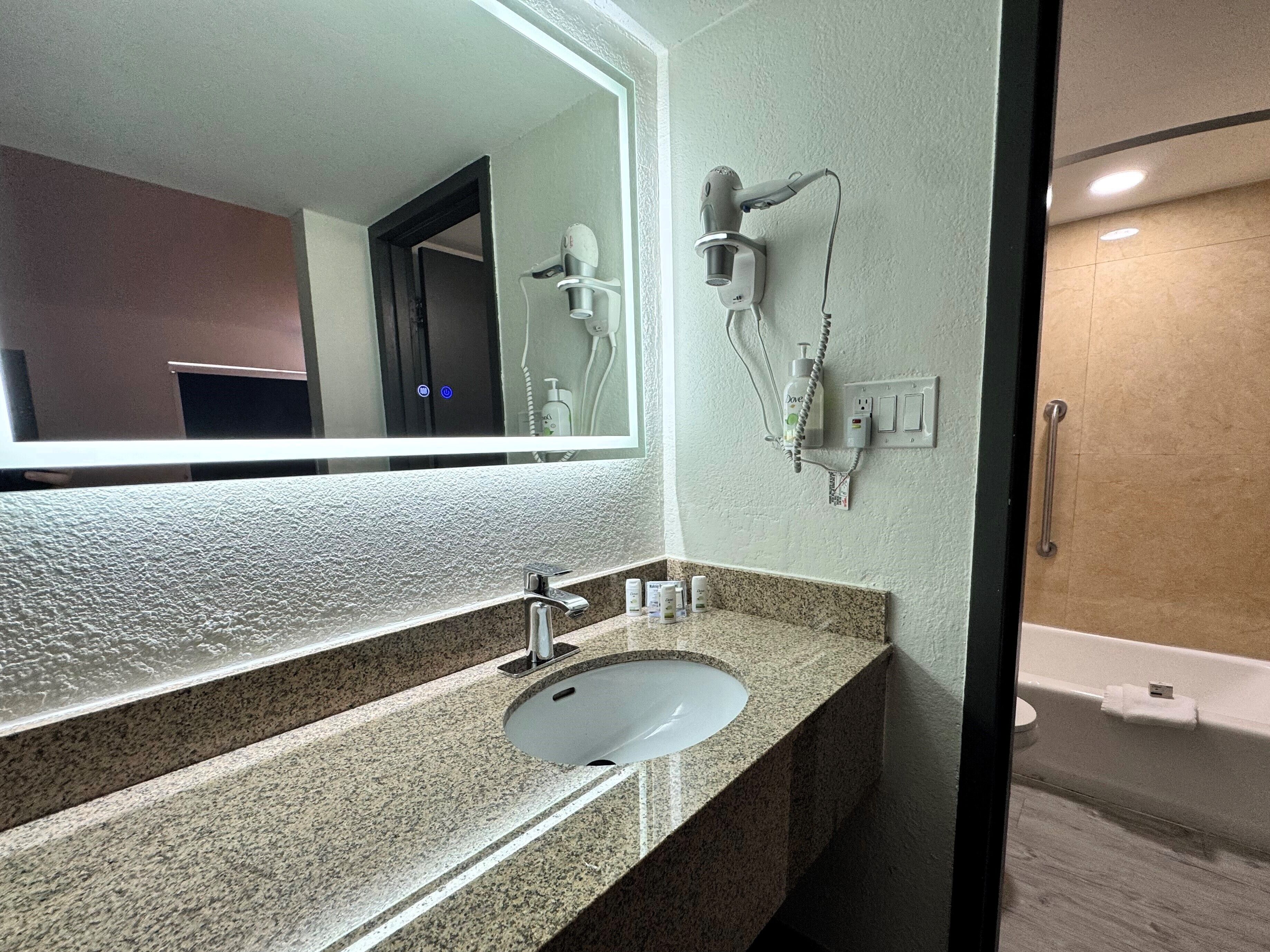 A bathroom with a granite countertop, a white sink, a large mirror, a hairdryer, and a shower.