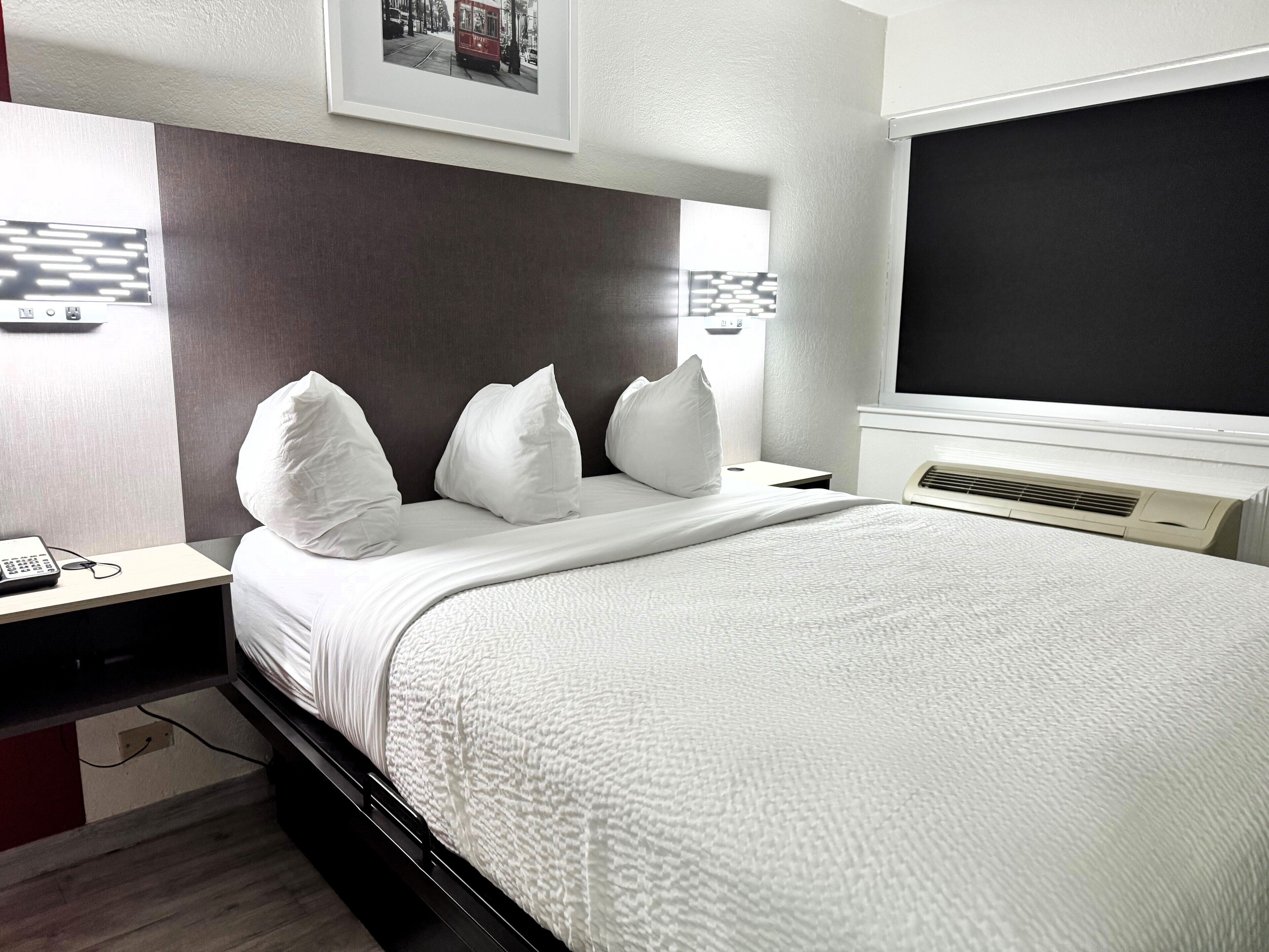A modern hotel room with a large bed, white bedding, a headboard, a bedside table with a phone, and a wall-mounted TV.