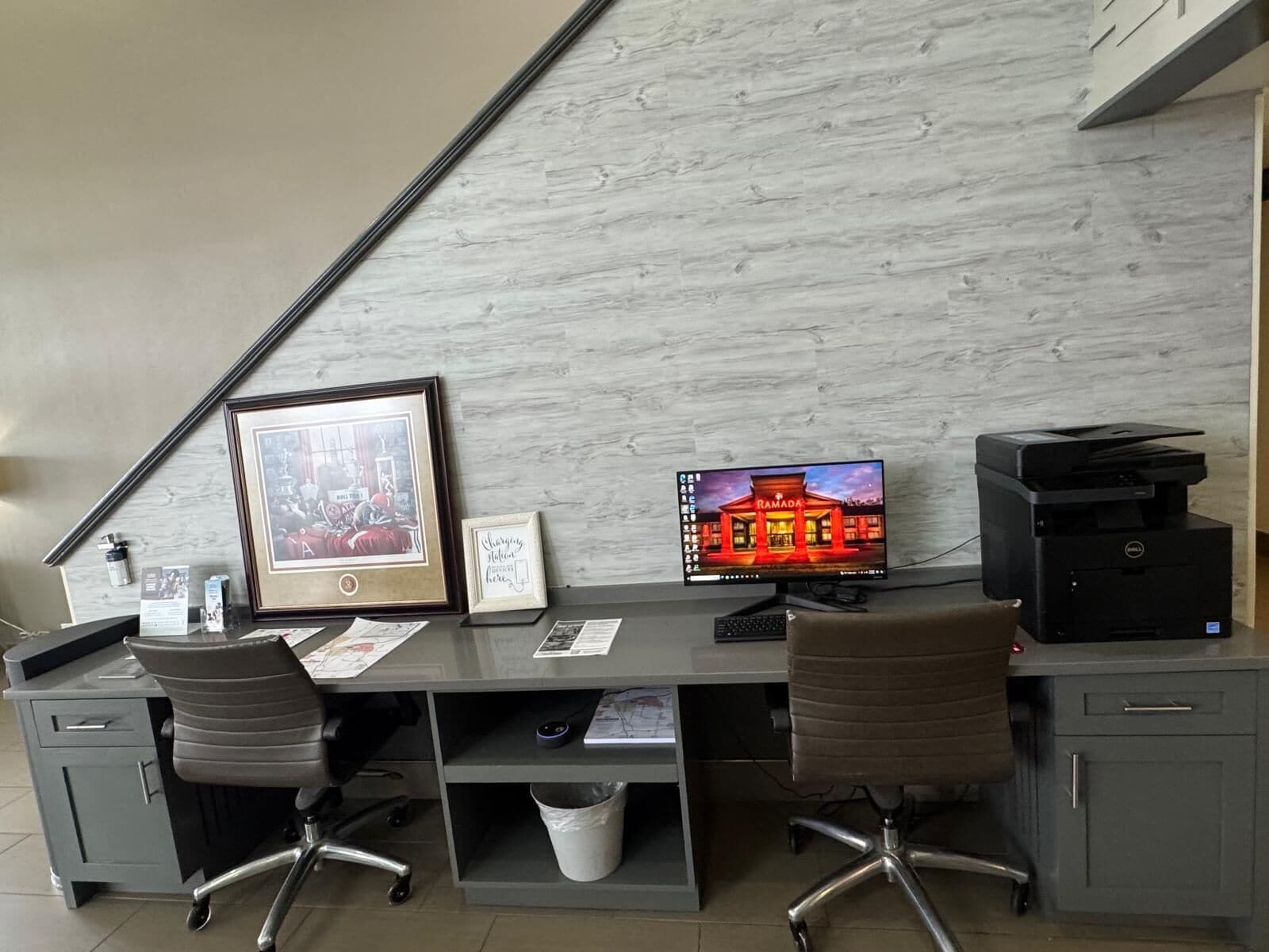A desk with two office chairs, a computer monitor displaying a Ramadan-themed image, a printer, and framed pictures on the desk.