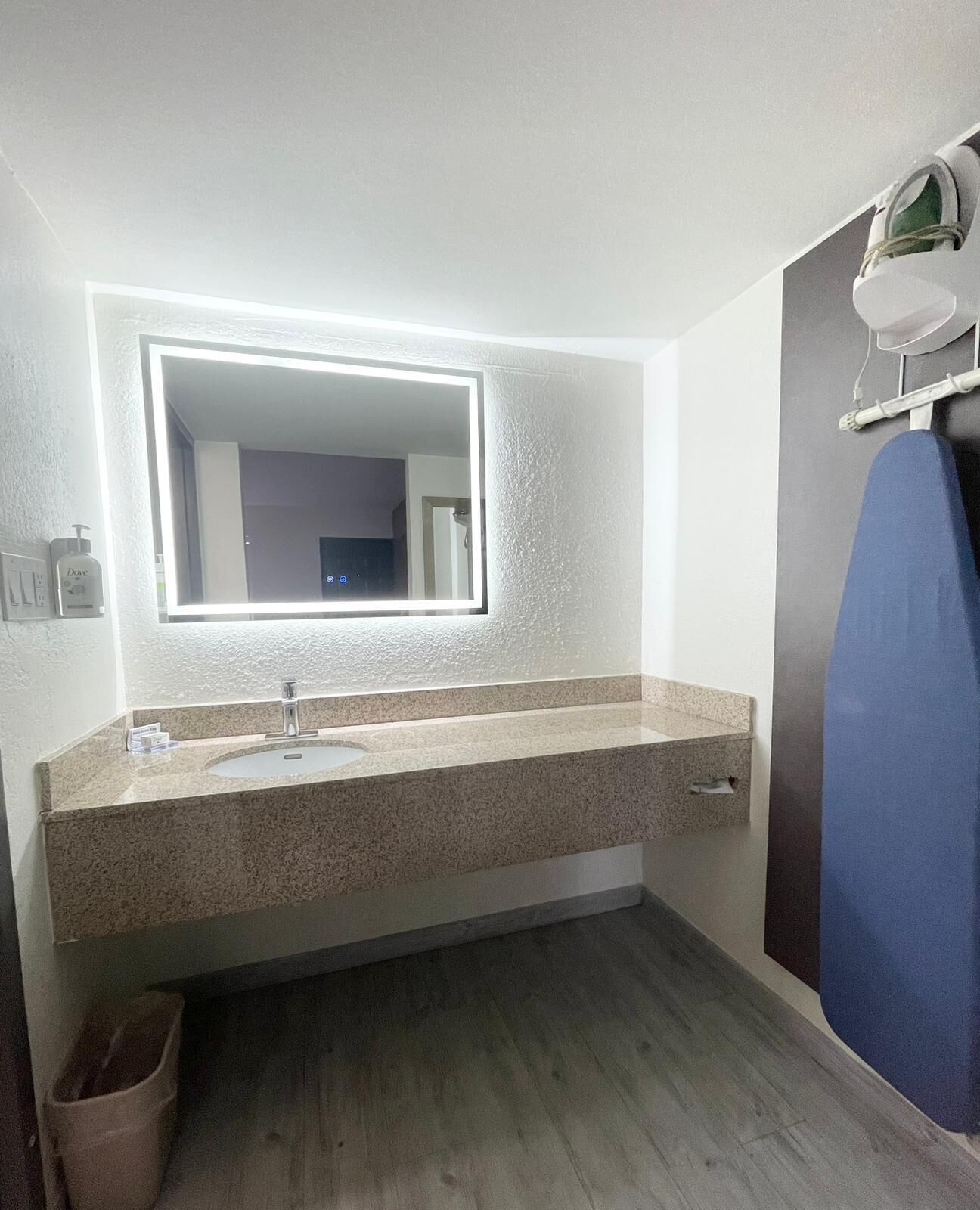 A bathroom with a granite countertop sink, a mirror with integrated lighting, a blue towel rack, and a trash bin.