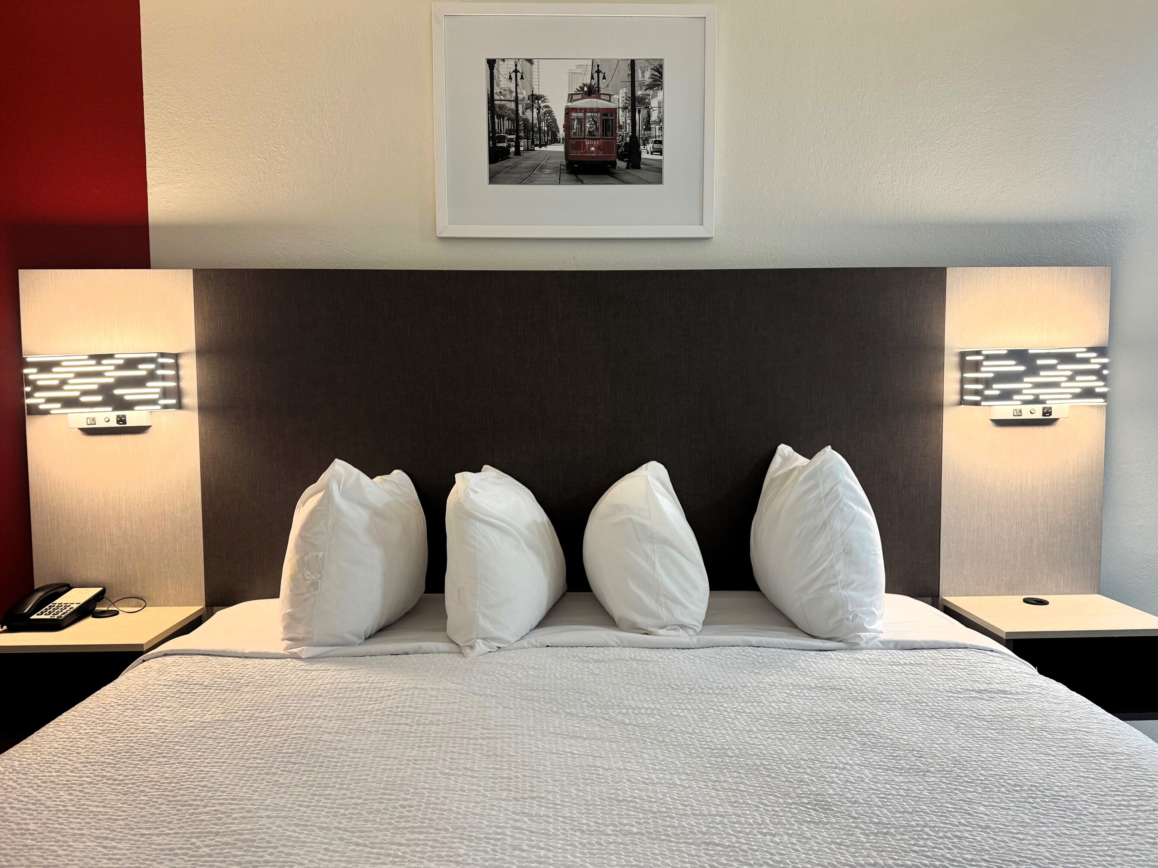 Una habitación de hotel con una cama grande, almohadas blancas, un cuadro enmarcado sobre la cama y lámparas de noche.
