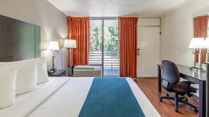 Une chambre d’hôtel avec un lit, un bureau, une chaise, une lampe et une fenêtre avec des rideaux.