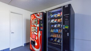 Un distributeur automatique arborant le design de Coca-Cola et doté d’un panneau de commande à boutons-poussoirs, situé dans un couloir avec une porte blanche et un sol carrelé.