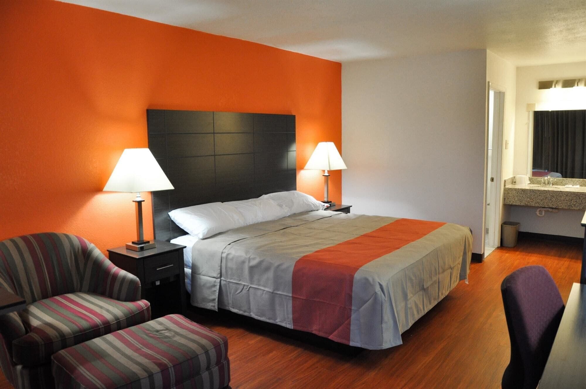 Habitación de hotel con una cama, dos lámparas de noche, un sillón a rayas, un reposapiés a juego, y un baño visible a través de una puerta abierta.