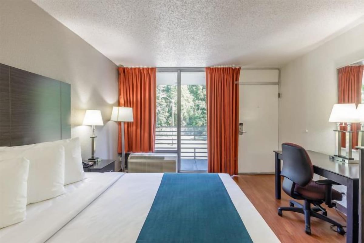 A hotel room with a bed, desk, chair, and a view of the outdoors through a sliding door.