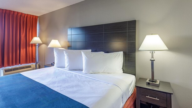 Une chambre d’hôtel avec un grand lit, deux tables de chevet avec des lampes, un radiateur et une fenêtre avec des rideaux rouges.