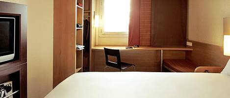 Una habitación de hotel con cama, escritorio, silla, televisor y ventana con cortinas.