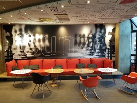 Un área de descanso moderna con un mural grande en la pared, asientos rojos y mesas redondas.