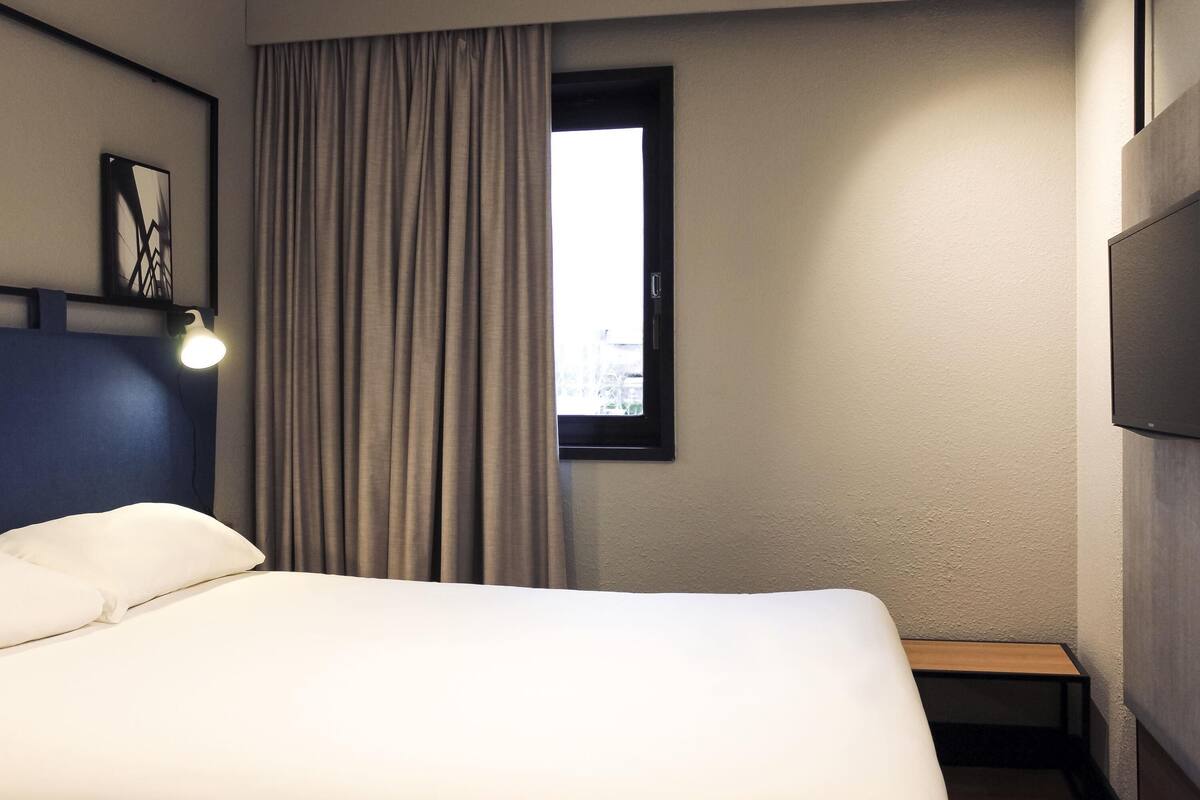 Ein Hotelzimmer mit Bett, Nachttischlampe, einem Fenster mit Vorhängen, einem Wand-Fernseher und einem kleinen Tisch.
