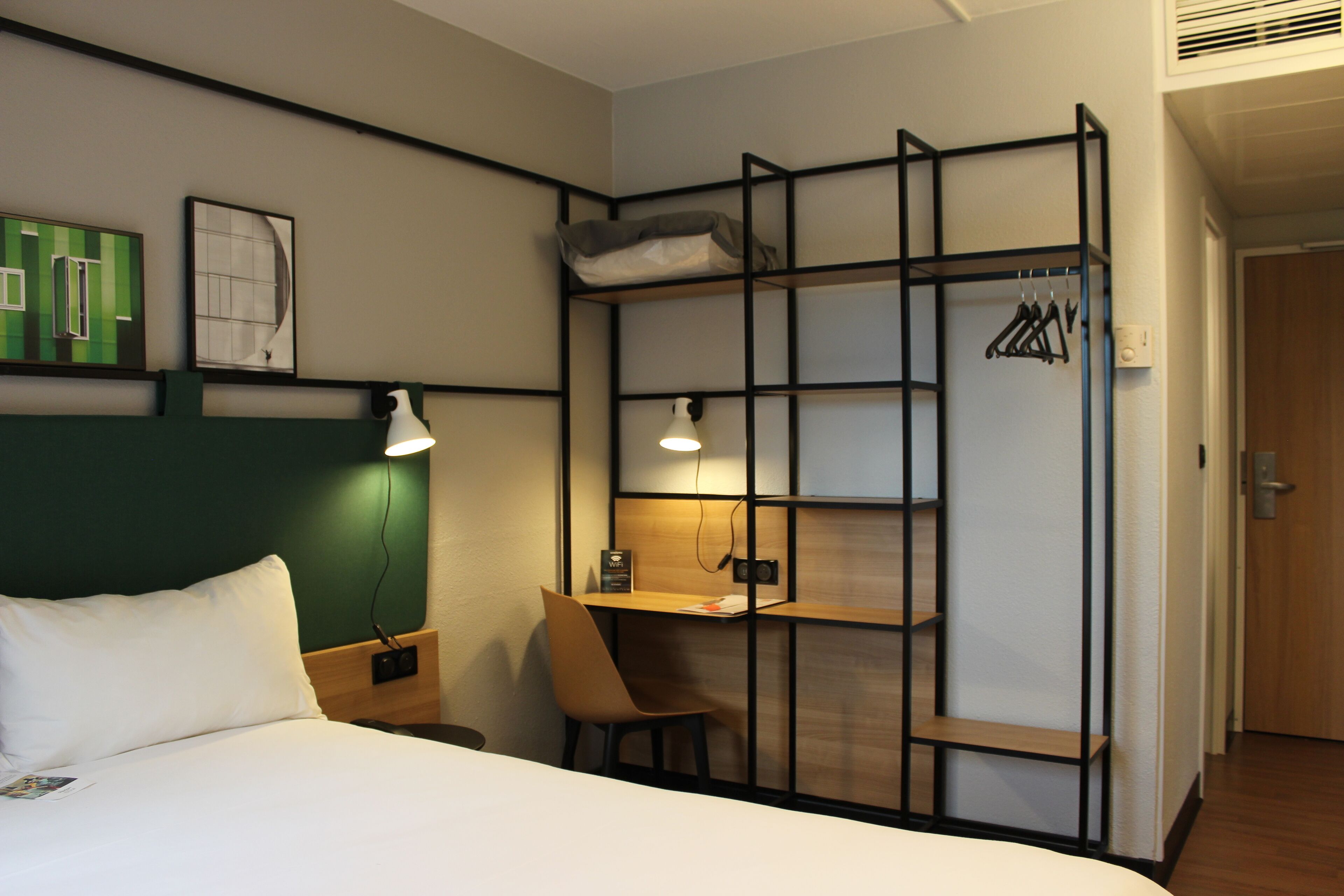 A hotel room with a bed, a desk with a chair, a bookshelf, and a coat rack.