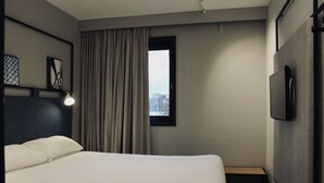 Una camera d'albergo con un letto, una finestra con tende, un comodino e una TV a parete.