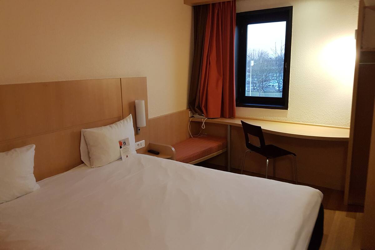 Ein Hotelzimmer mit Bett, Schreibtisch, Stuhl, einem Fenster mit Vorhängen und einer Wandleuchte.