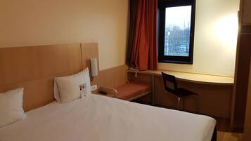 Une chambre d’hôtel comprenant un lit, un bureau, une chaise, une fenêtre avec des rideaux et un luminaire fixé au mur.