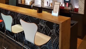 Un'area bar con un bancone in legno, sedie bianche e una parete con motivi astratti.