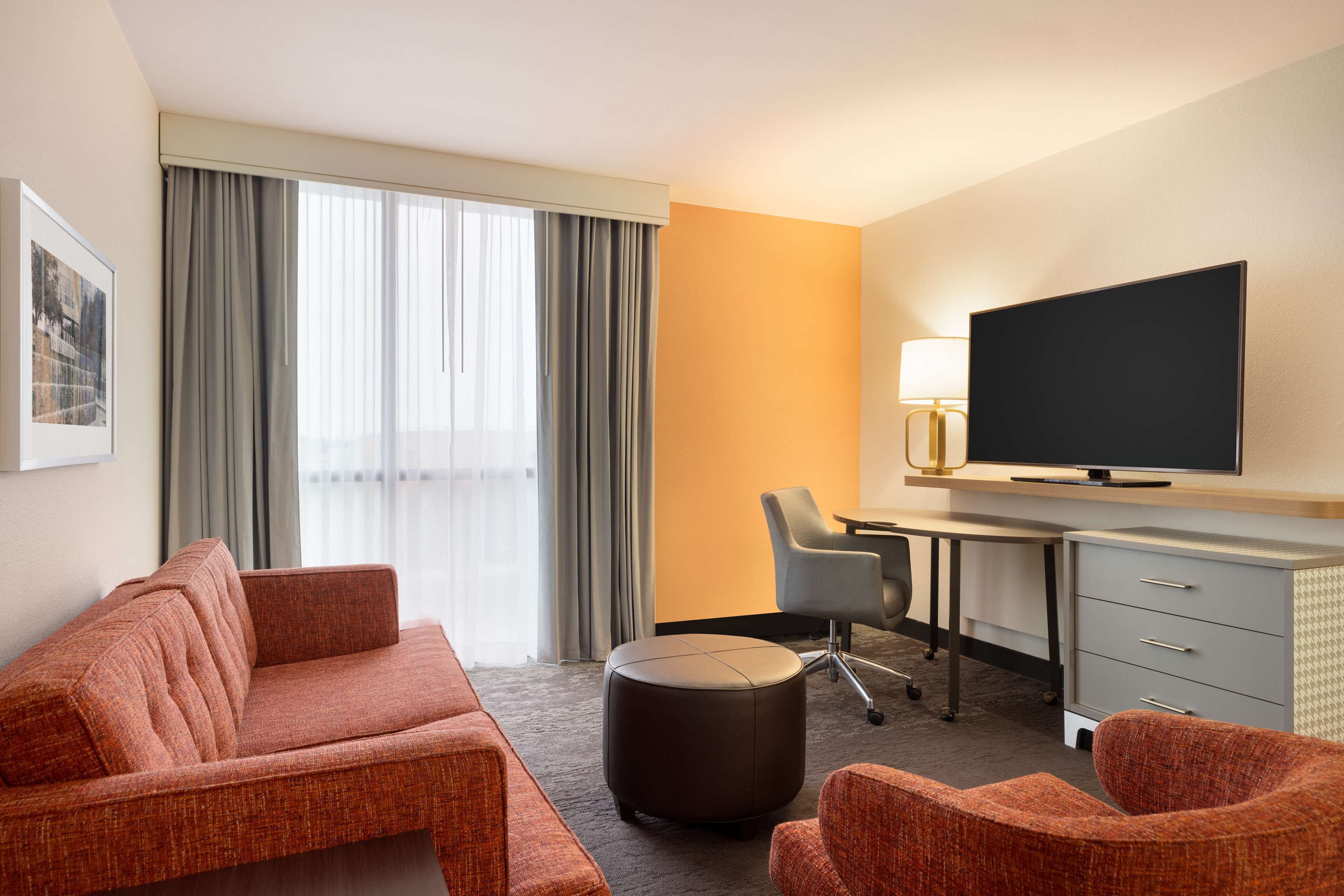 A hotel room with a sofa, ottoman, desk, chair, TV, and a large window with curtains.