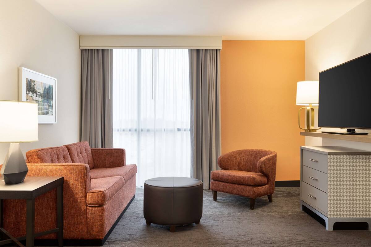 A hotel room with a sofa, armchair, ottoman, television, and a desk with a lamp.