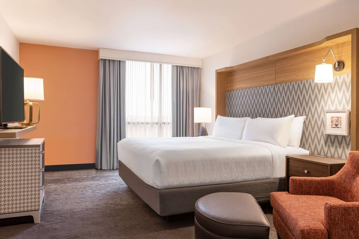 A hotel room with a large bed, a desk with a lamp, a chair, and a TV.
