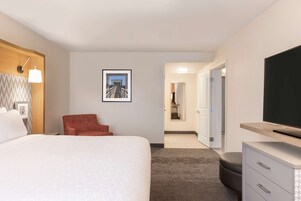Habitación de hotel con una cama grande, un sillón rojo, un televisor de pantalla plana, un escritorio con cajones y un cuadro en la pared.