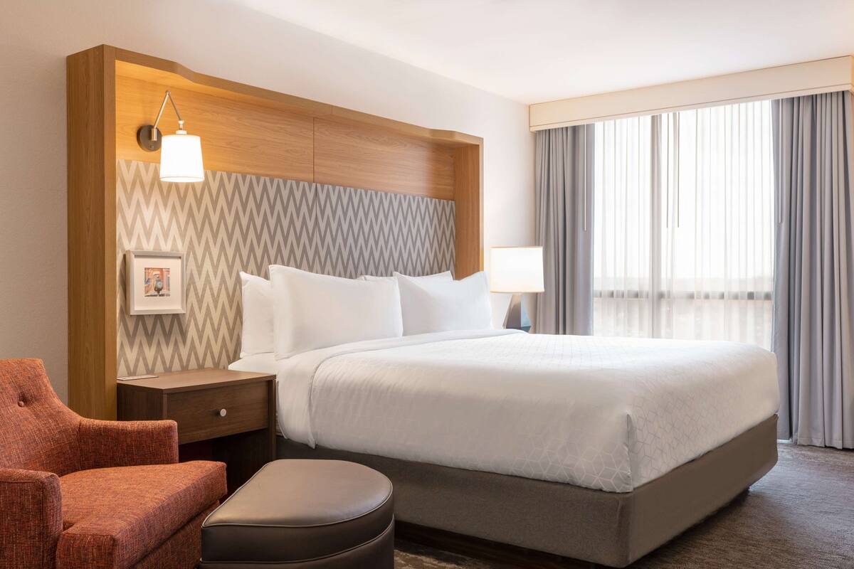 A hotel room with a large bed, a wooden headboard, a nightstand, a chair, and a window with curtains.