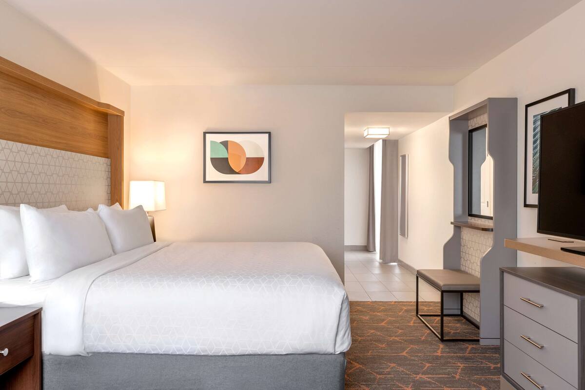 A hotel room with a bed, bedside table, lamp, TV, and a desk with drawers.