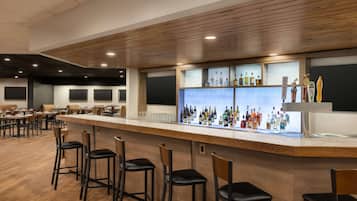 Eine Bar mit Holzhockern, einer langen Theke mit verschiedenen Flaschen und Gläsern sowie einer Sitzgelegenheit mit Tischen und Stühlen.