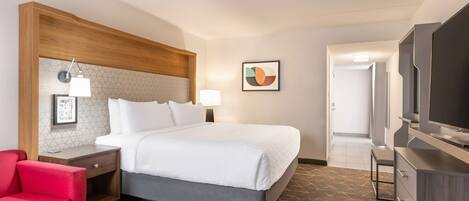 A hotel room with a bed, a red chair, a desk with a lamp, a television, and a framed picture on the wall.