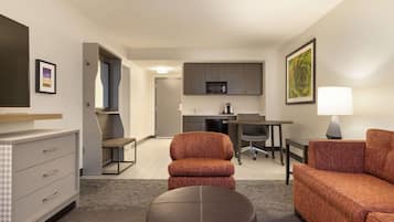 Una habitación de hotel moderna con sofá, reposapiés y escritorio con silla. Cuenta con una kitchenette con microondas y una mesa de comedor.