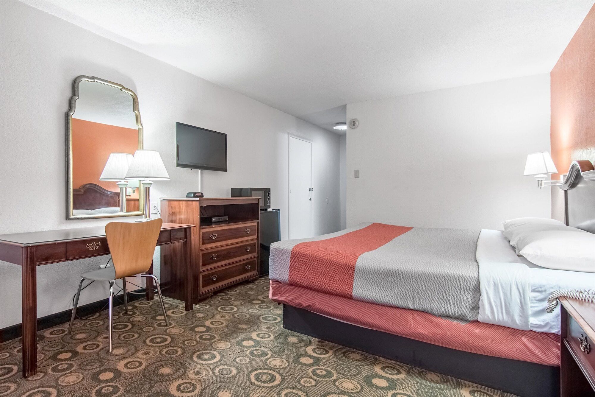 A hotel room with a bed, a desk with a chair, a dresser, a television, and a patterned carpet.