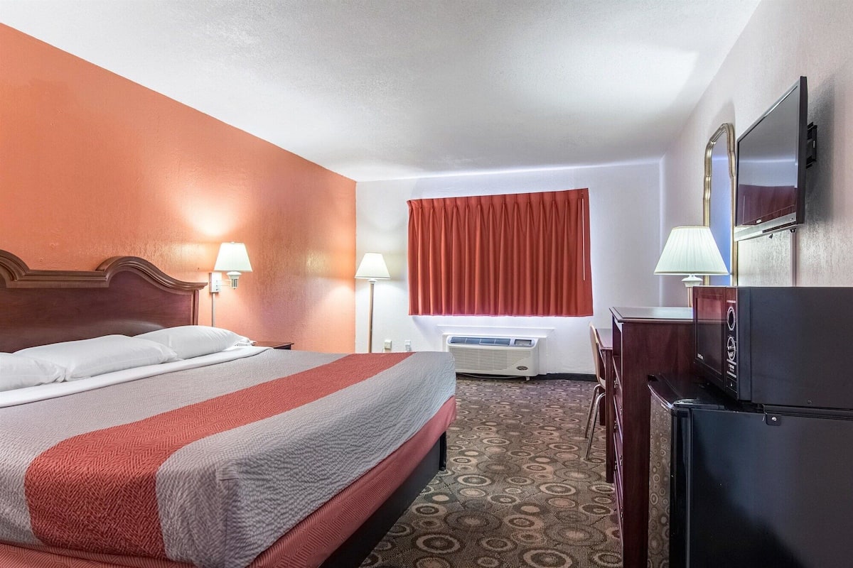 A hotel room with a bed, a television, a desk, a lamp, and an air conditioning unit.