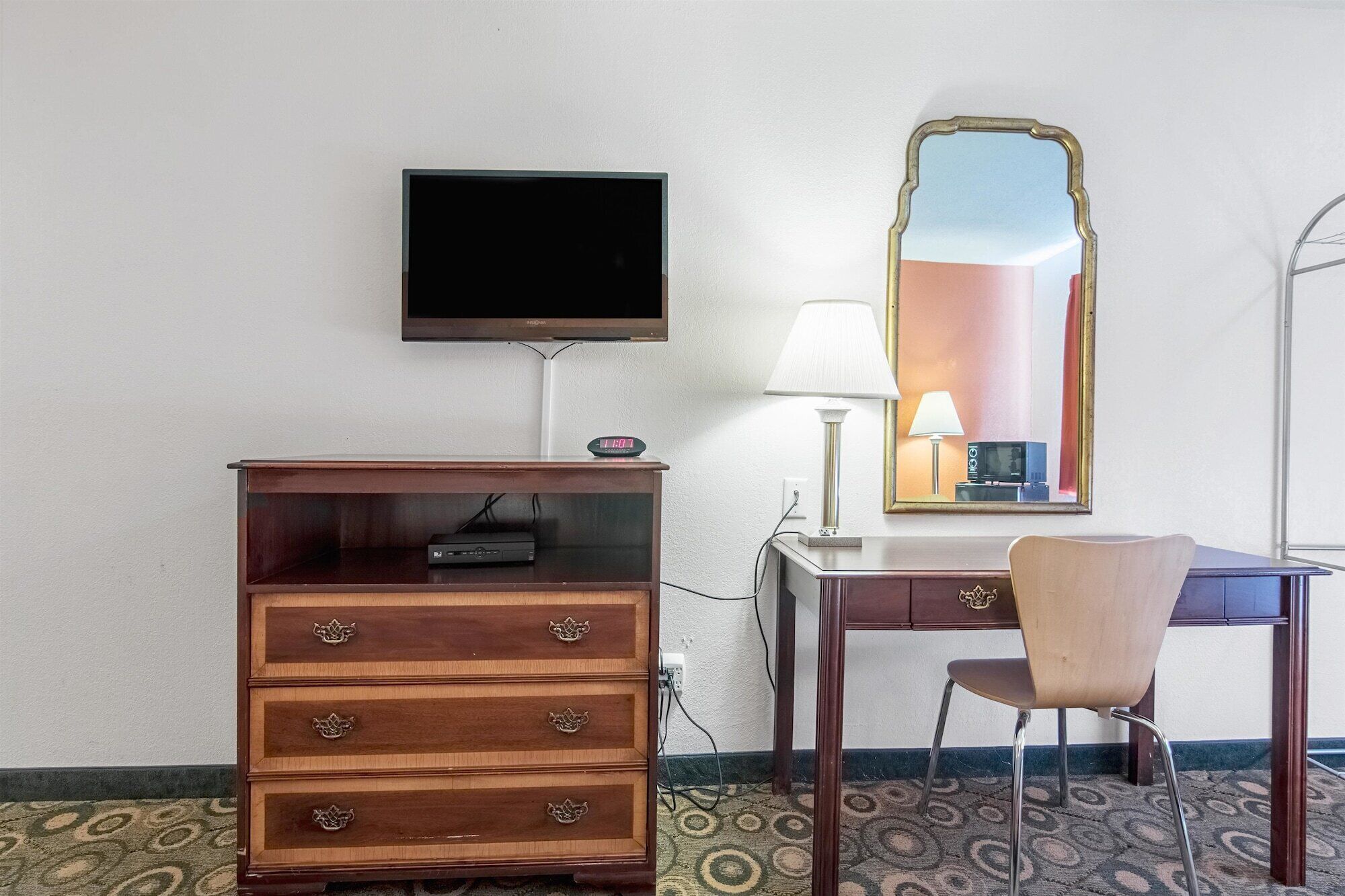 A hotel room with a wooden dresser, a television mounted on the wall, a desk with a chair, a mirror, and a lamp.