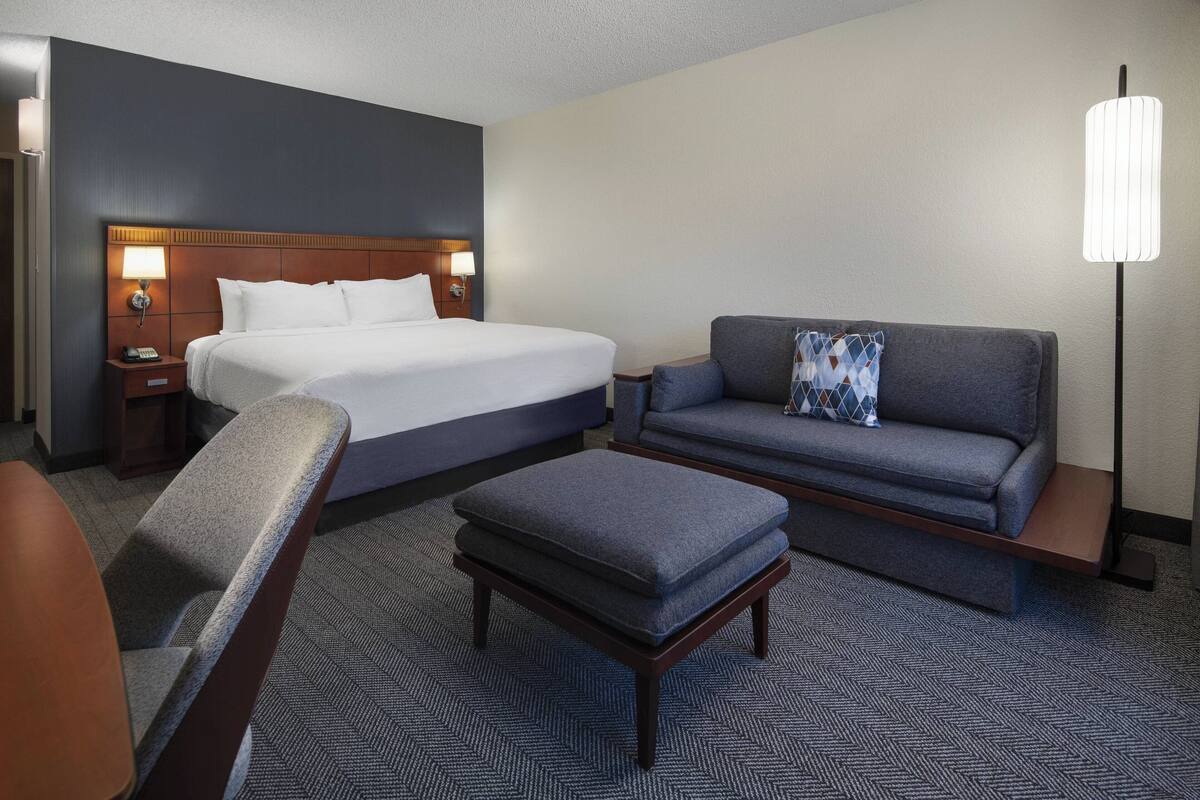 Ein Hotelzimmer mit einem großen Bett, einer Couch, einem Hocker und einem Schreibtisch mit Lampe.