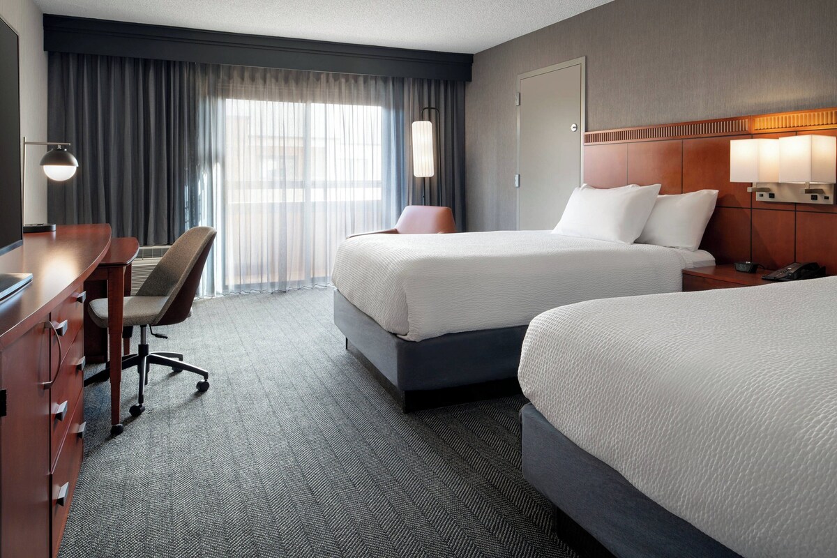 A hotel room with two beds, a desk, a chair, and a lamp.