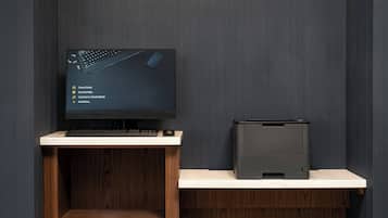 A modern workspace with a flat-screen monitor, keyboard, and printer on a wooden desk against a dark wall.